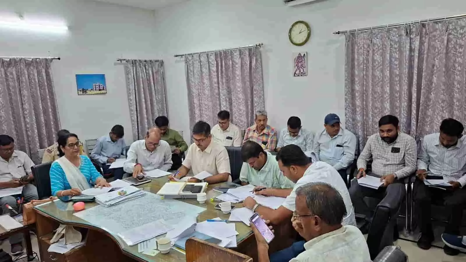 तस्वीर में मीटिंग हॉल में मेज के चारों ओर बैठे अधिकारी कागजात देखते हुए। तस्वीर में मीटिंग हॉल में मेज के चारों ओर बैठे अधिकारी कागजात देखते हुए।