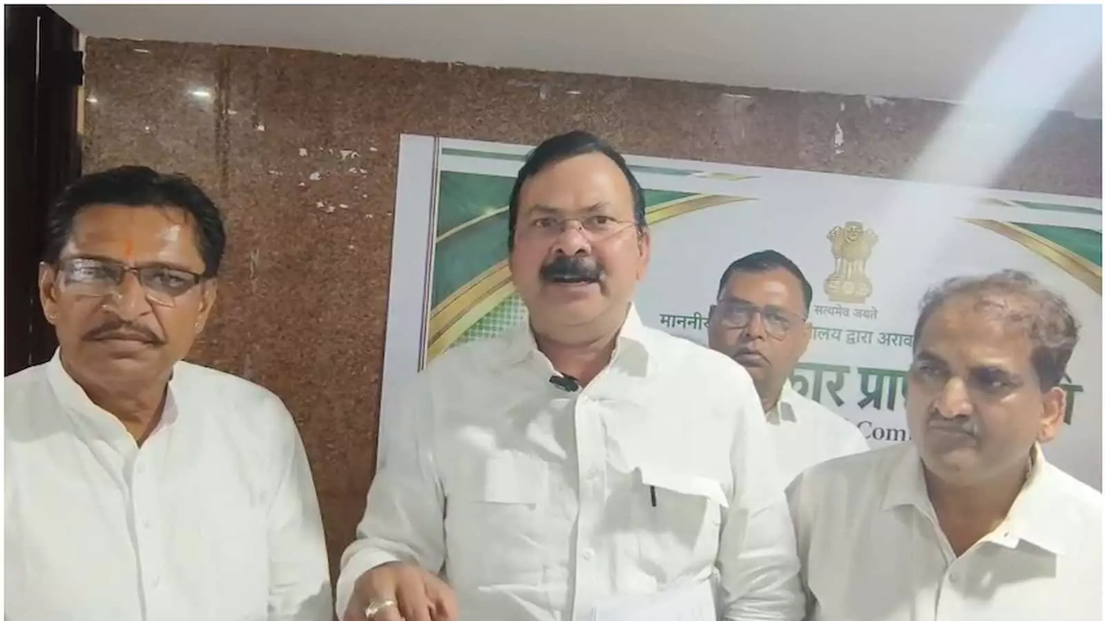 तस्वीर में चार पुरुष खड़े हैं, बीच का व्यक्ति हाथ से इशारा कर रहा है, पृष्ठभूमि में बोर्ड है। तस्वीर में चार पुरुष खड़े हैं, बीच का व्यक्ति हाथ से इशारा कर रहा है, पृष्ठभूमि में बोर्ड है।