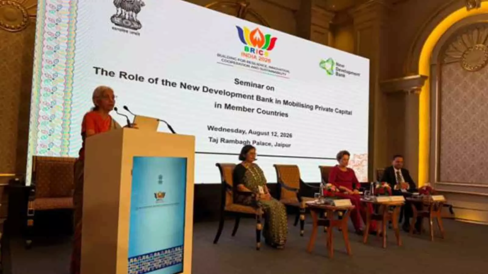 मंच पर पोडियम से भाषण देतीं वित्त मंत्री और बैठे हुए अन्य अधिकारी जयपुर में। मंच पर पोडियम से भाषण देतीं वित्त मंत्री और बैठे हुए अन्य अधिकारी जयपुर में।