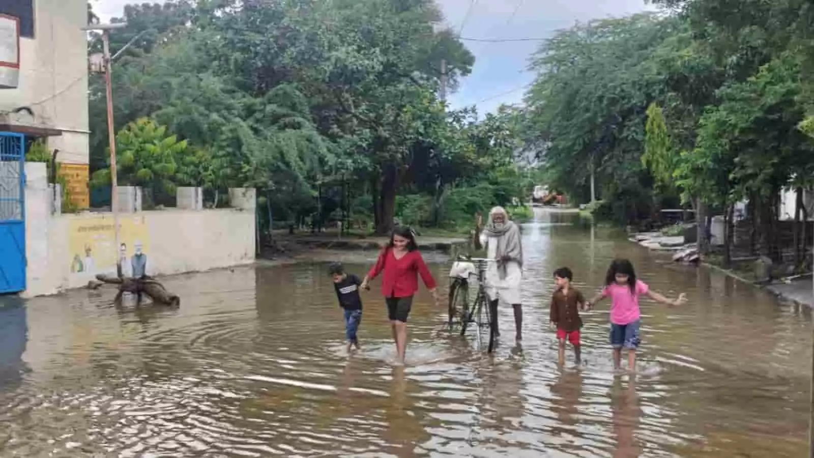 तस्वीर में पानी से भरी सड़क पर बच्चे चलते और खेलते हुए दिखाई दे रहे हैं। तस्वीर में पानी से भरी सड़क पर बच्चे चलते और खेलते हुए दिखाई दे रहे हैं।