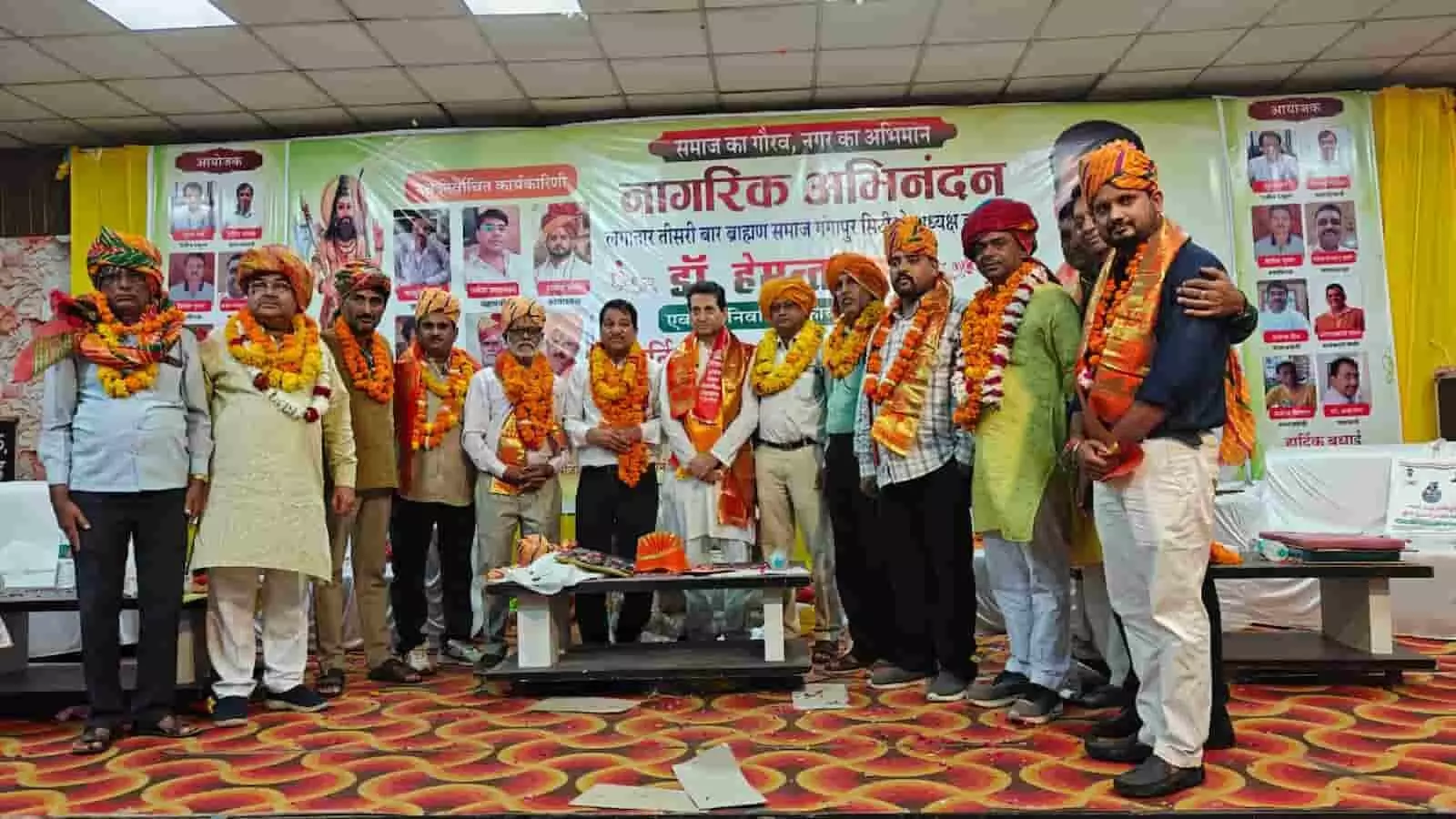 मंच पर साफा और माला पहने कई पुरुष एक समारोह में खड़े हैं। मंच पर साफा और माला पहने कई पुरुष एक समारोह में खड़े हैं।