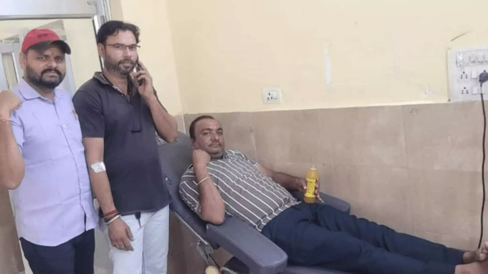 रक्तदान कक्ष में तीन पुरुष, एक कुर्सी पर रक्तदान करते हुए रक्तदान कक्ष में तीन पुरुष, एक कुर्सी पर रक्तदान करते हुए