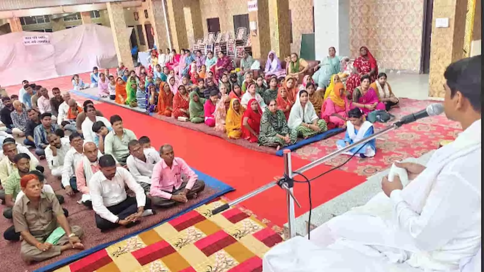 सफेद वस्त्र पहने वक्ता माइक्रोफोन से संबोधित करते हुए, सभागार में श्रोता बैठे हैं। सफेद वस्त्र पहने वक्ता माइक्रोफोन से संबोधित करते हुए, सभागार में श्रोता बैठे हैं।