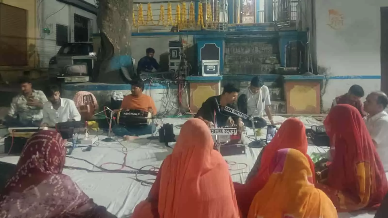 तस्वीर में मंदिर परिसर में भजन संध्या के दौरान प्रस्तुति देते गायक और बैठे श्रद्धालु दिख रहे हैं। तस्वीर में मंदिर परिसर में भजन संध्या के दौरान प्रस्तुति देते गायक और बैठे श्रद्धालु दिख रहे हैं।