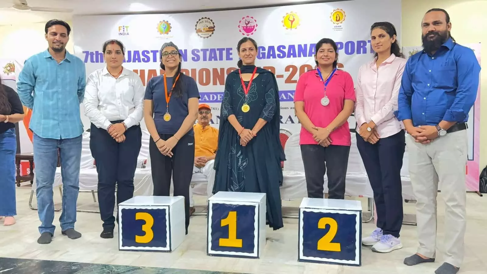 योगासन प्रतियोगिता के विजेता पोडियम पर मेडल पहनकर खड़े हुए खिलाड़ी और आयोजक योगासन प्रतियोगिता के विजेता पोडियम पर मेडल पहनकर खड़े हुए खिलाड़ी और आयोजक