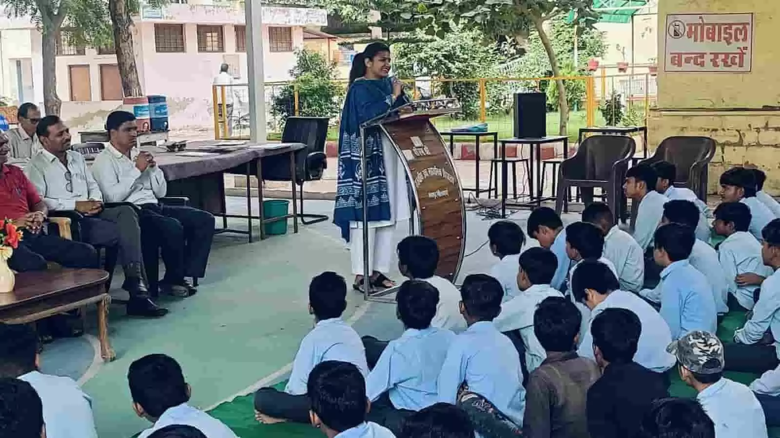 स्कूल परिसर में छात्रों को संबोधित करती महिला न्यायाधीश और मंच पर बैठे अतिथि। स्कूल परिसर में छात्रों को संबोधित करती महिला न्यायाधीश और मंच पर बैठे अतिथि।