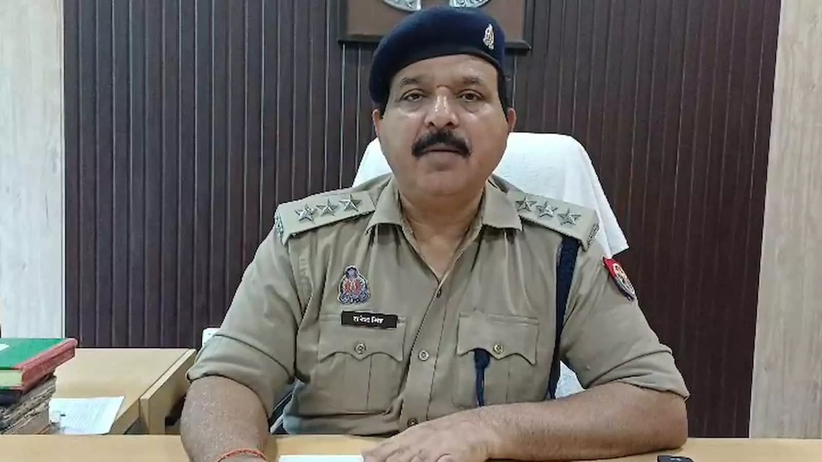 तस्वीर में एक पुलिस अधिकारी पुलिस की वर्दी पहनकर कार्यालय में मेज के पास बैठे हैं। तस्वीर में एक पुलिस अधिकारी पुलिस की वर्दी पहनकर कार्यालय में मेज के पास बैठे हैं।