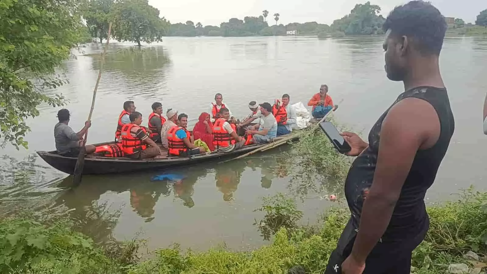 तस्वीर में बाईं ओर लाइफ जैकेट पहने लोग नाव में हैं और दाईं ओर एक व्यक्ति खड़ा है।