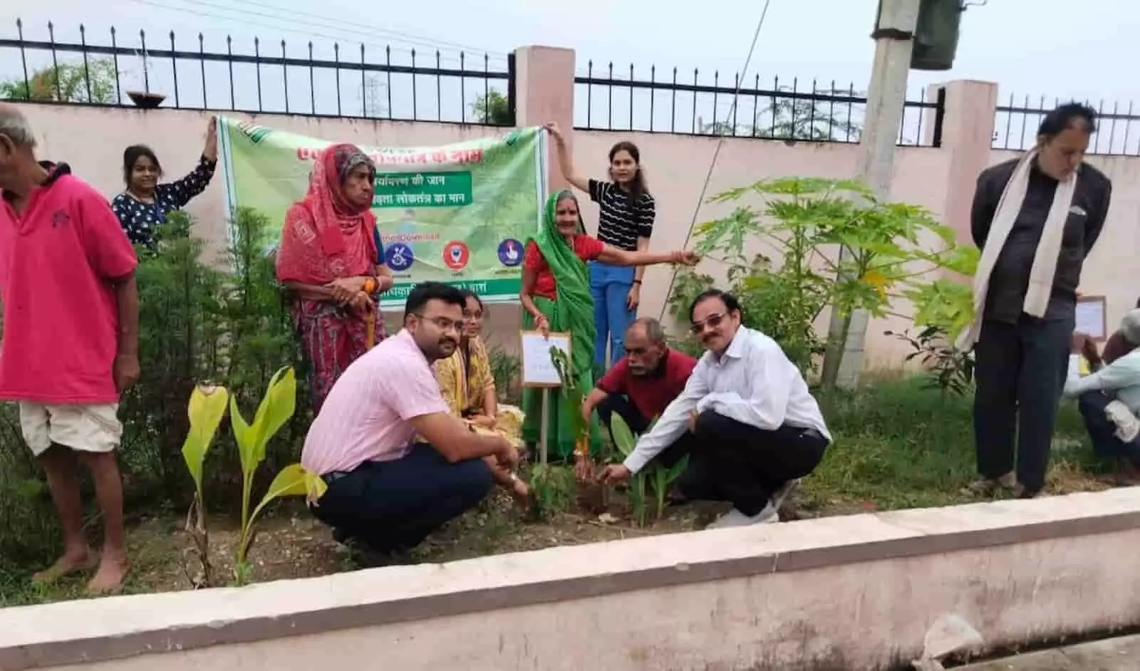 तस्वीर में बाएं से दाएं महिलाएं और पुरुष बैनर पकड़े हुए बगीचे में पौधारोपण करते दिख रहे हैं। तस्वीर में बाएं से दाएं महिलाएं और पुरुष बैनर पकड़े हुए बगीचे में पौधारोपण करते दिख रहे हैं।
