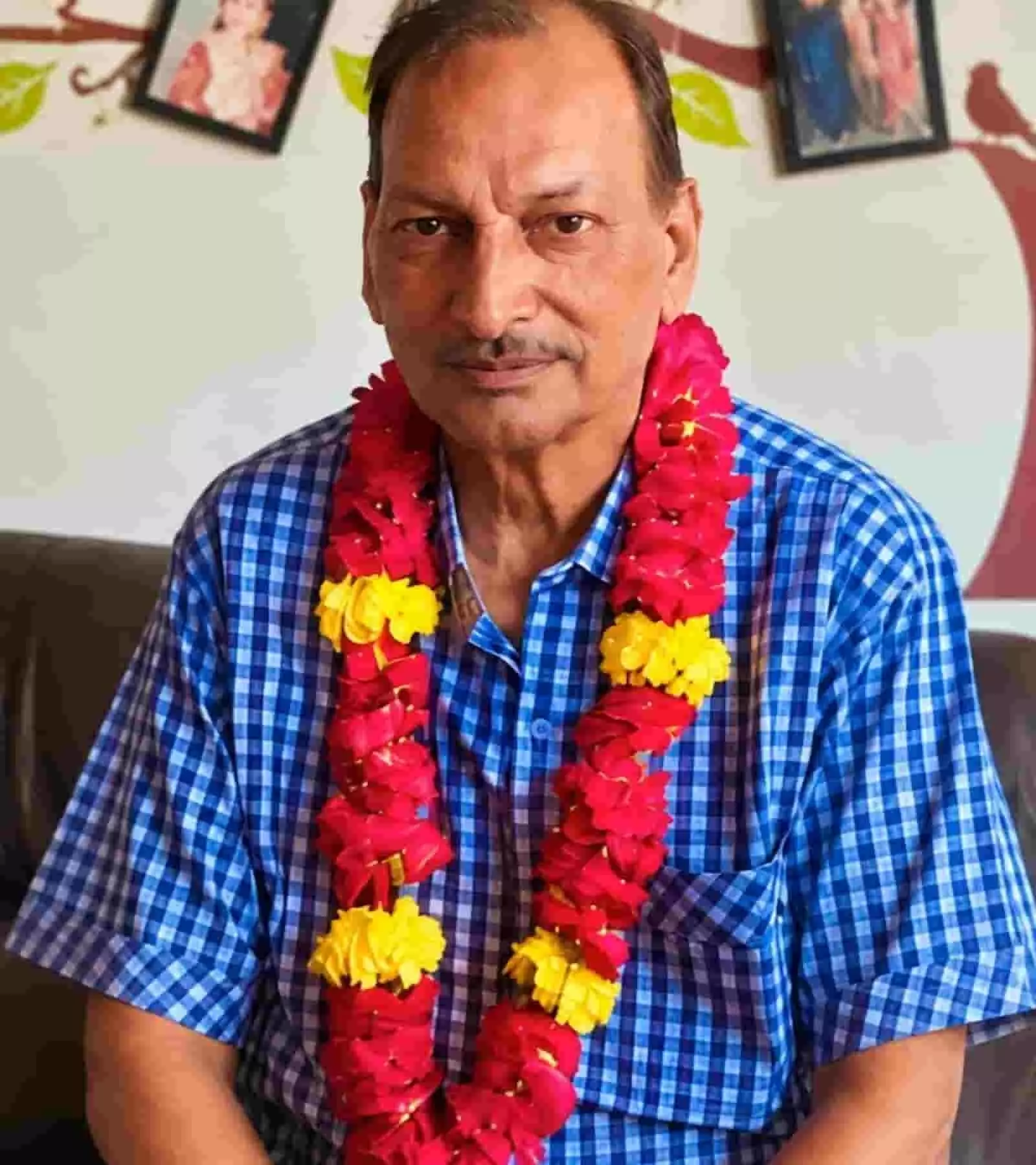 तस्वीर में दिख रही दीवार के आगे नीली चेकदार शर्ट और माला पहने व्यक्ति बैठे हैं। तस्वीर में दिख रही दीवार के आगे नीली चेकदार शर्ट और माला पहने व्यक्ति बैठे हैं।