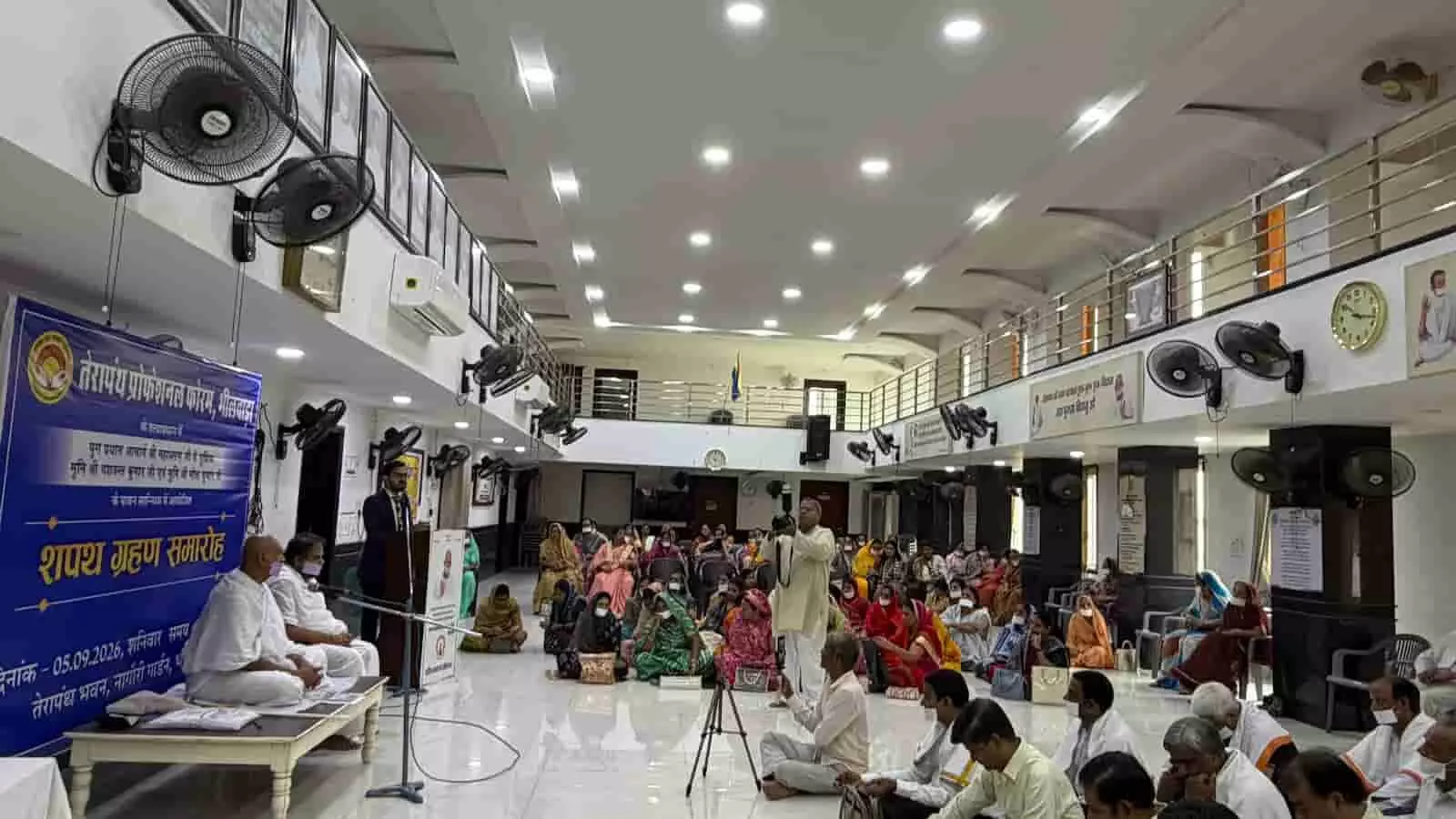 हॉल में बाएं ओर श्वेत वस्त्रधारी साधु मंच पर बैठे हैं और सामने लोग उपस्थित हैं। हॉल में बाएं ओर श्वेत वस्त्रधारी साधु मंच पर बैठे हैं और सामने लोग उपस्थित हैं।
