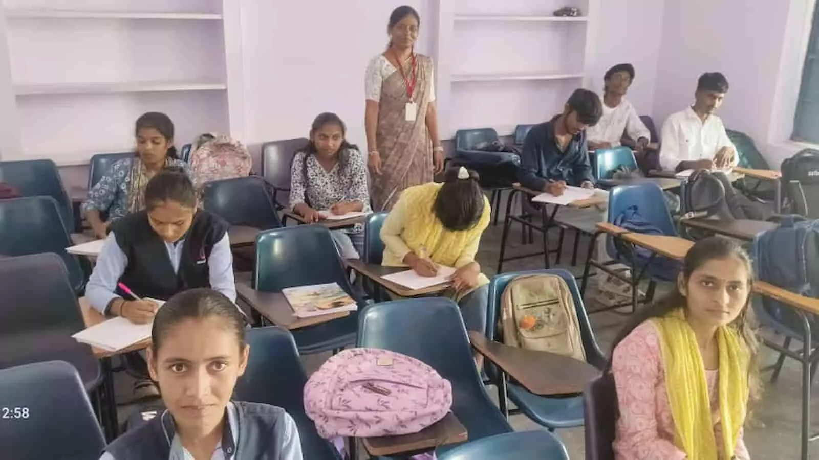 तस्वीर में दिख रही कक्षा में छात्राएं और छात्र कुर्सियों पर बैठकर निबंध प्रतियोगिता में लिख रहे हैं।