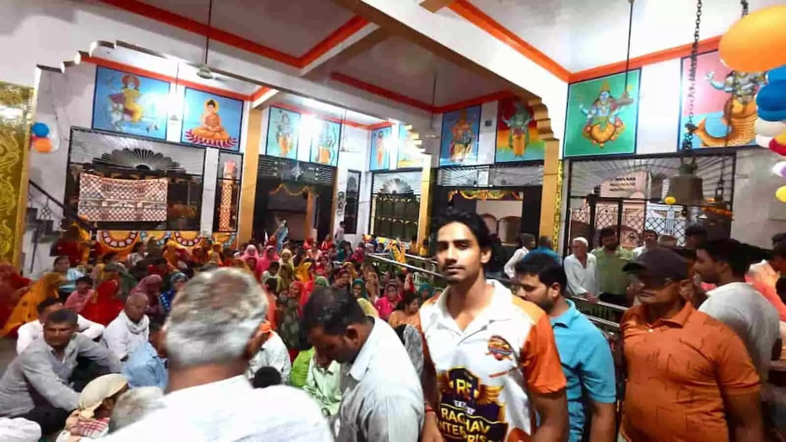 तस्वीर में मंदिर हॉल के भीतर श्रद्धालु फर्श पर बैठे हैं और पुरुष सामने खड़े दिख रहे हैं। तस्वीर में मंदिर हॉल के भीतर श्रद्धालु फर्श पर बैठे हैं और पुरुष सामने खड़े दिख रहे हैं।