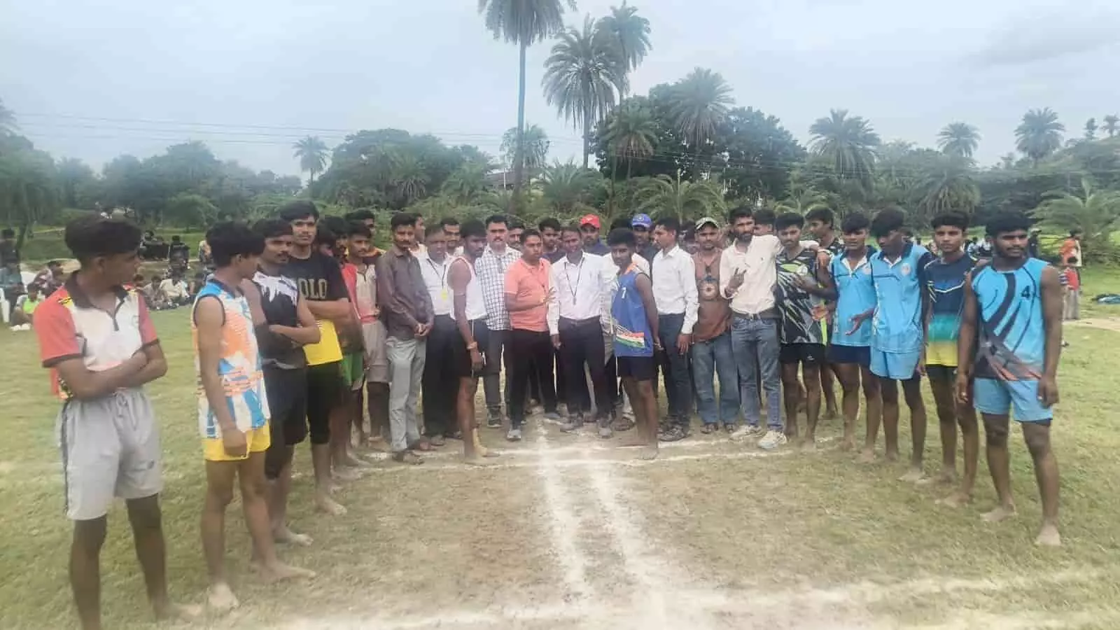 तस्वीर में खेल के मैदान पर सफेद रेखाओं के पीछे युवा खिलाड़ी और आयोजक खड़े हैं। तस्वीर में खेल के मैदान पर सफेद रेखाओं के पीछे युवा खिलाड़ी और आयोजक खड़े हैं।
