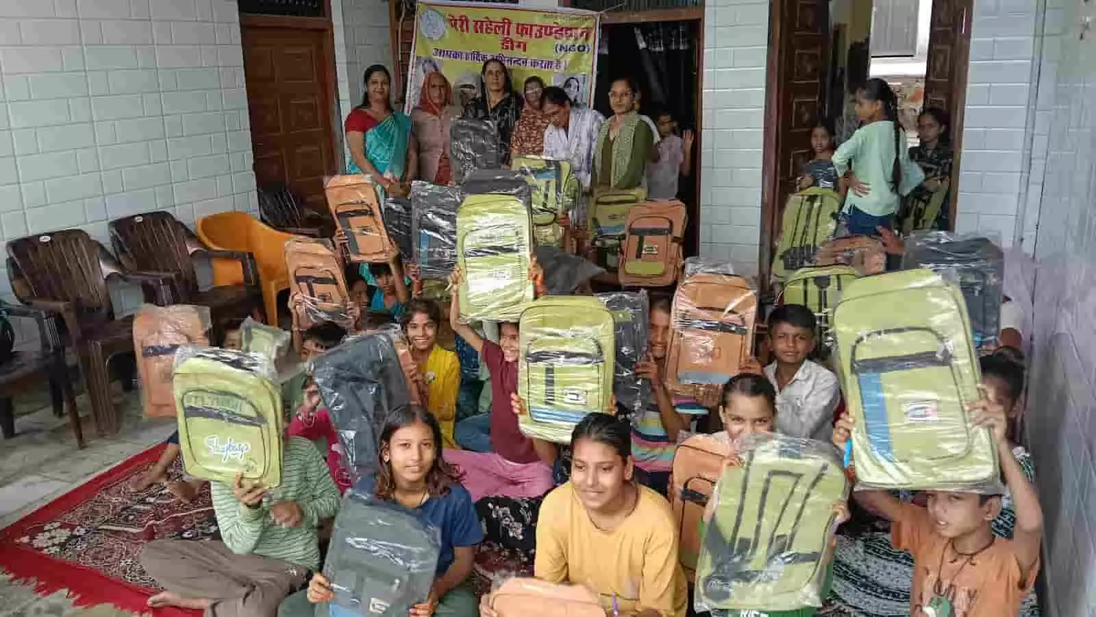 तस्वीर में बच्चे हाथ में स्कूल बैग लेकर एक बैनर के सामने बैठे हुए हैं। तस्वीर में बच्चे हाथ में स्कूल बैग लेकर एक बैनर के सामने बैठे हुए हैं।