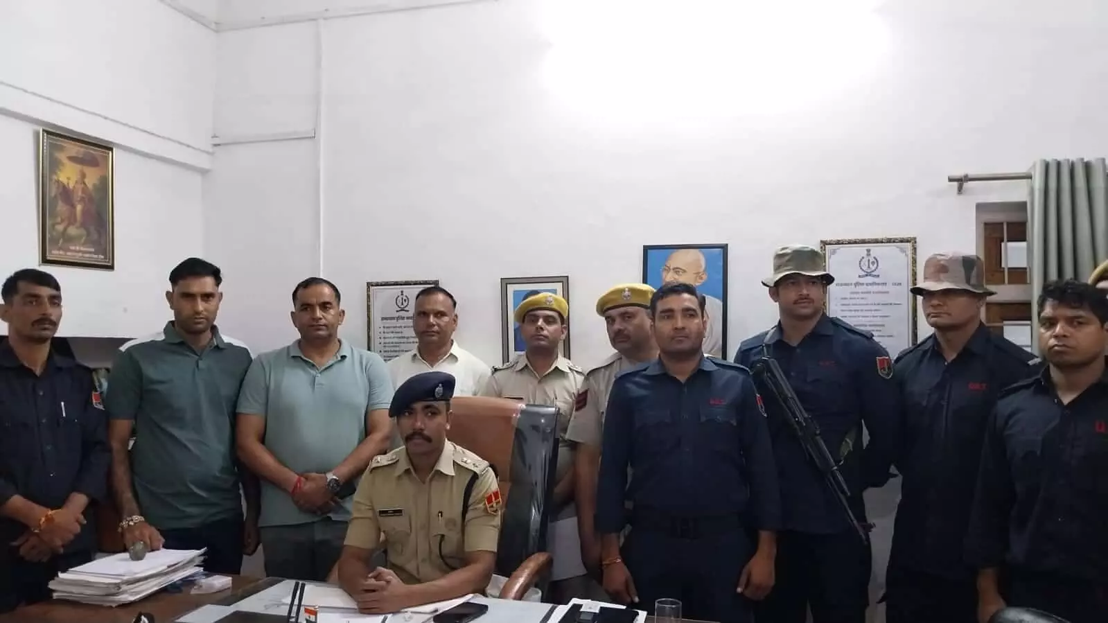 तस्वीर में वर्दीधारी पुलिसकर्मी और सादे कपड़ों में लोग ऑफिस की मेज के पीछे खड़े हैं। तस्वीर में वर्दीधारी पुलिसकर्मी और सादे कपड़ों में लोग ऑफिस की मेज के पीछे खड़े हैं।