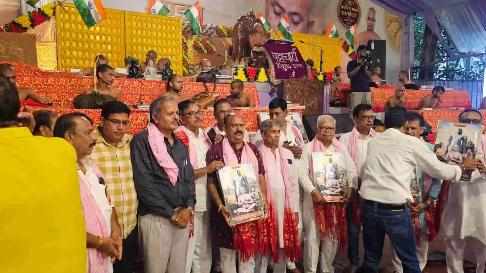 तस्वीर में पुरुष हाथ में चित्र पकड़े मंच के सामने खड़े हैं और पीछे साधु बैठे हैं। तस्वीर में पुरुष हाथ में चित्र पकड़े मंच के सामने खड़े हैं और पीछे साधु बैठे हैं।