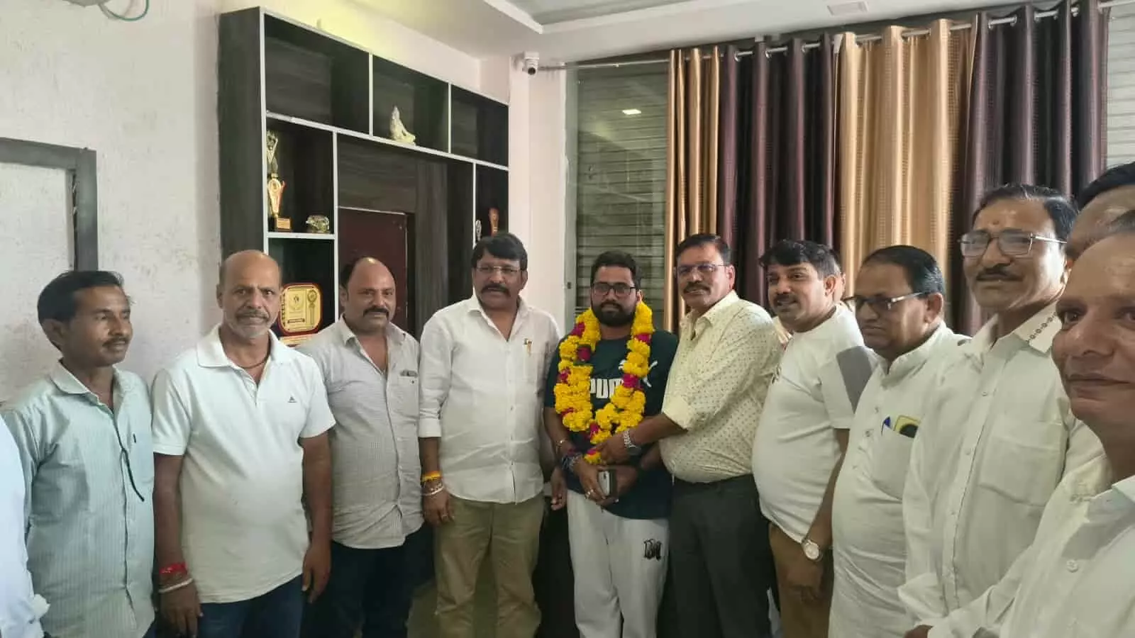तस्वीर में कमरे के भीतर खड़े पुरुष और बीच में पीली माला पहना हुआ एक व्यक्ति खड़ा। तस्वीर में कमरे के भीतर खड़े पुरुष और बीच में पीली माला पहना हुआ एक व्यक्ति खड़ा।