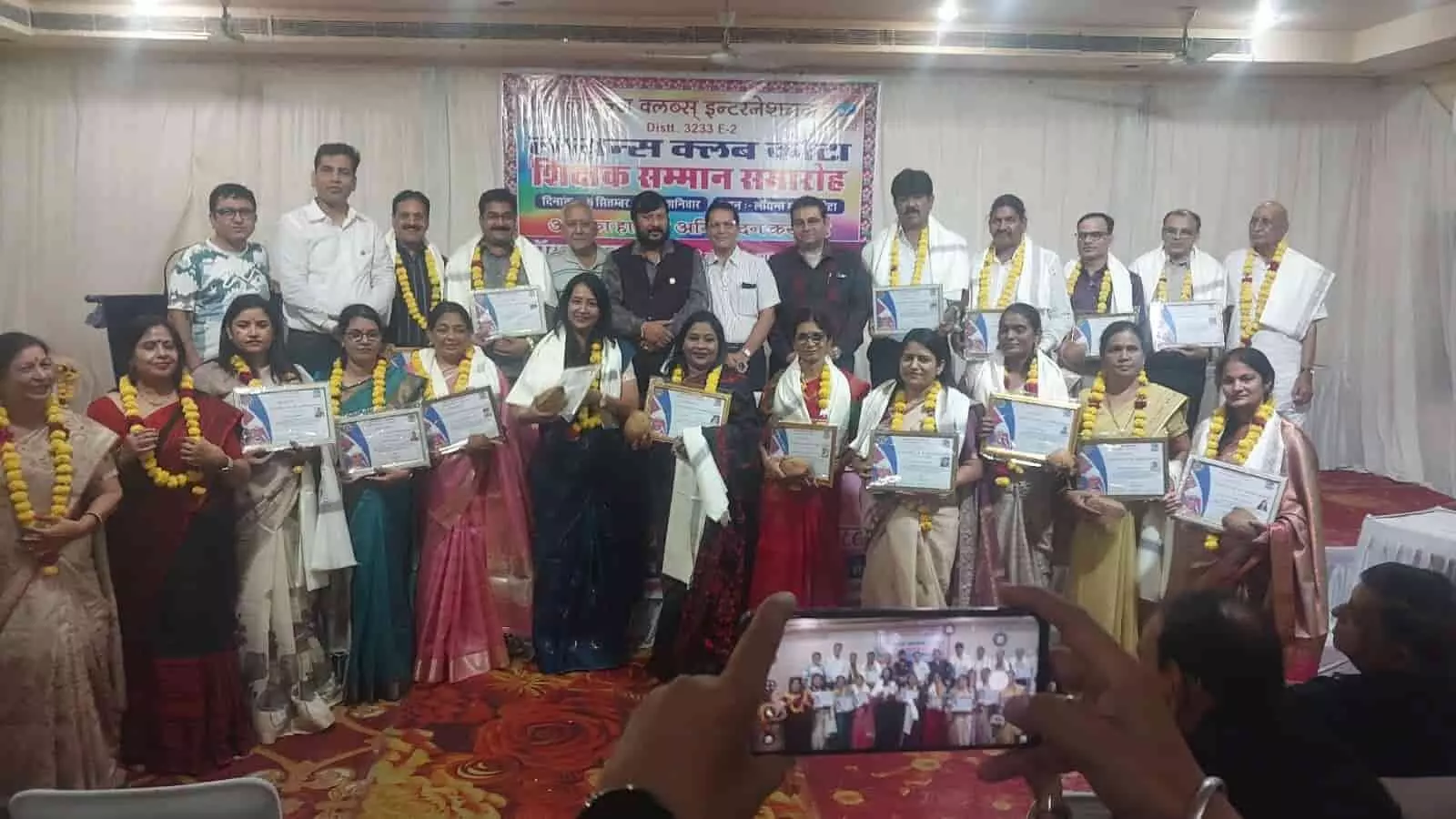 तस्वीर में लायंस क्लब के बैनर के सामने पुरुष और महिला शिक्षक पुरस्कार पकड़े खड़े हैं। तस्वीर में लायंस क्लब के बैनर के सामने पुरुष और महिला शिक्षक पुरस्कार पकड़े खड़े हैं।