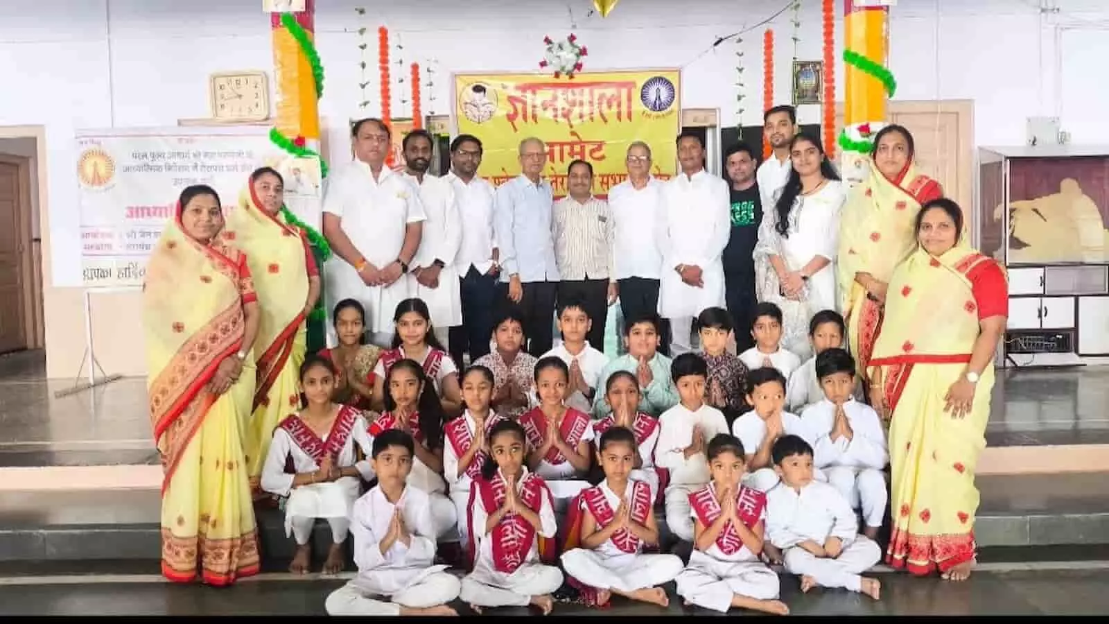 तस्वीर में बैनर के सामने वयस्क खड़े हैं और आगे बच्चे प्रार्थना की मुद्रा में बैठे हैं। तस्वीर में बैनर के सामने वयस्क खड़े हैं और आगे बच्चे प्रार्थना की मुद्रा में बैठे हैं।