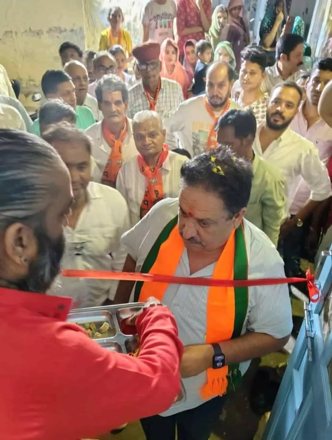 तस्वीर में लाल शर्ट वाला व्यक्ति ट्रे पकड़े है और भगवा दुपट्टा पहने व्यक्ति रिबन काट रहा है। तस्वीर में लाल शर्ट वाला व्यक्ति ट्रे पकड़े है और भगवा दुपट्टा पहने व्यक्ति रिबन काट रहा है।