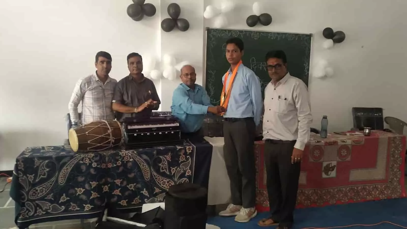 तस्वीर में दिख रही एक कमरे में बाएं से दाएं खड़े शिक्षक, छात्र को सम्मानित करते हुए। तस्वीर में दिख रही एक कमरे में बाएं से दाएं खड़े शिक्षक, छात्र को सम्मानित करते हुए।