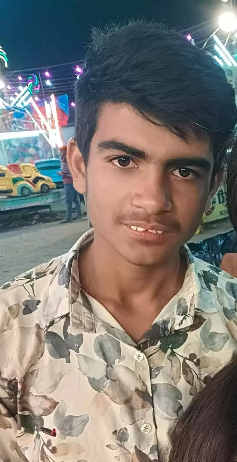 तस्वीर में प्रिंटेड शर्ट पहना एक किशोर मेले के झूलों और लाइटों के सामने खड़ा है। तस्वीर में प्रिंटेड शर्ट पहना एक किशोर मेले के झूलों और लाइटों के सामने खड़ा है।