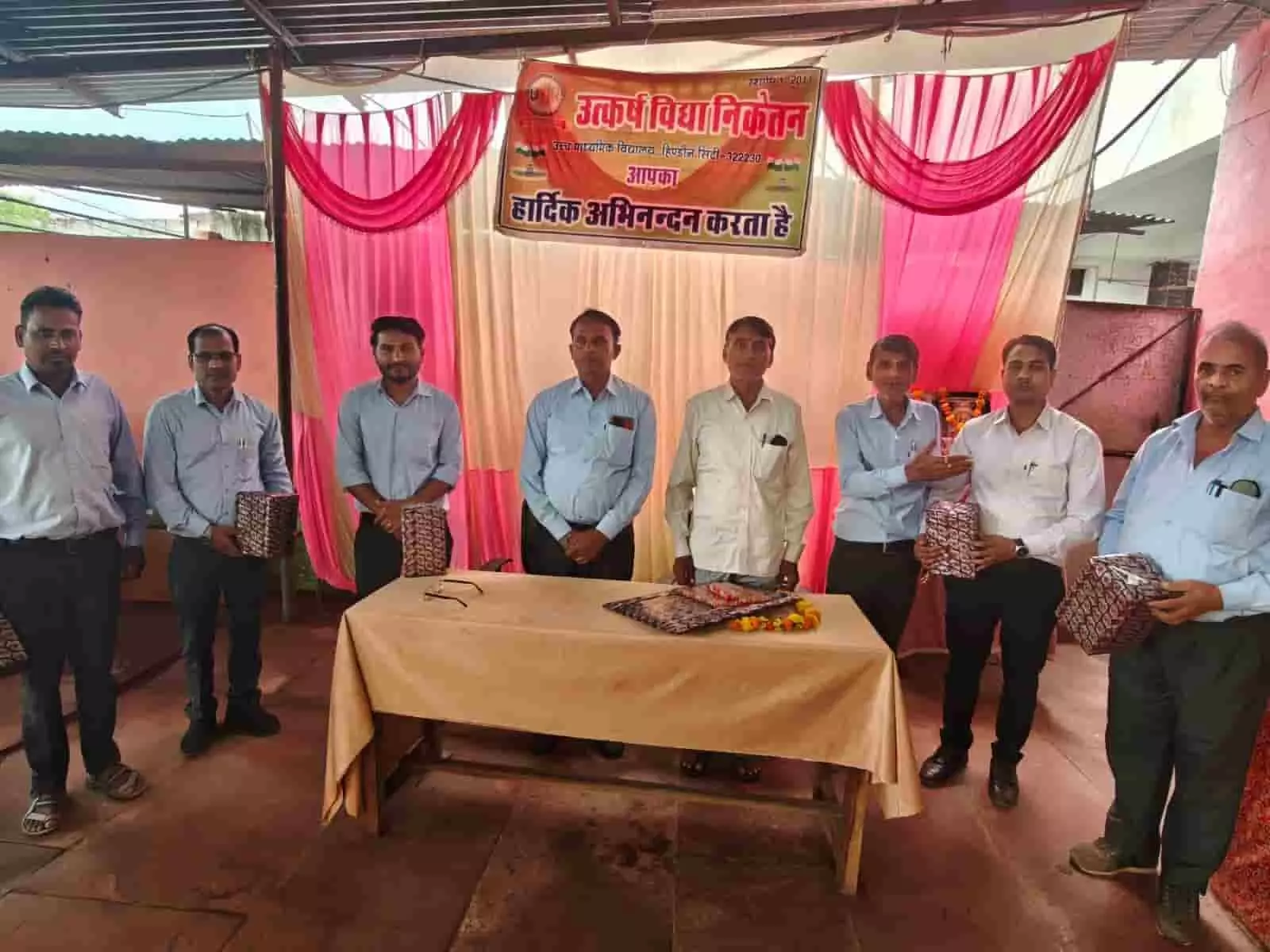 तस्वीर में दिख रही मेज के पीछे खड़े शिक्षक और अतिथि उपहार लेकर पोज दे रहे हैं। तस्वीर में दिख रही मेज के पीछे खड़े शिक्षक और अतिथि उपहार लेकर पोज दे रहे हैं।