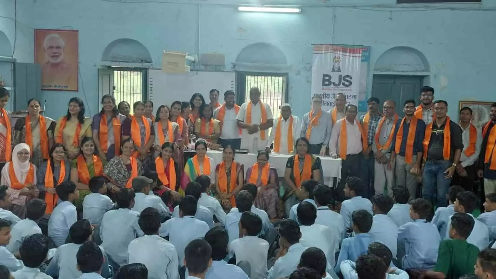 तस्वीर में बीजेएस सदस्य और शिक्षक नारंगी दुपट्टे पहनकर बैठे हुए स्कूली छात्रों के सामने खड़े हैं। तस्वीर में बीजेएस सदस्य और शिक्षक नारंगी दुपट्टे पहनकर बैठे हुए स्कूली छात्रों के सामने खड़े हैं।