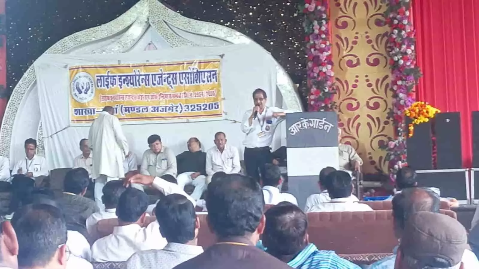 तस्वीर में दिख रहा वक्ता मंच पर बोल रहा है और सामने दर्शक बैठे हैं। तस्वीर में दिख रहा वक्ता मंच पर बोल रहा है और सामने दर्शक बैठे हैं।