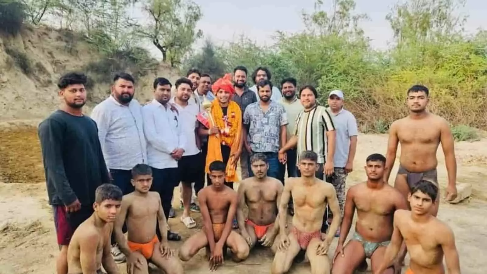 तस्वीर में दिख रहे पहलवान और पुरुष एक रेतीले मैदान में समूह बनाकर खड़े हैं। तस्वीर में दिख रहे पहलवान और पुरुष एक रेतीले मैदान में समूह बनाकर खड़े हैं।
