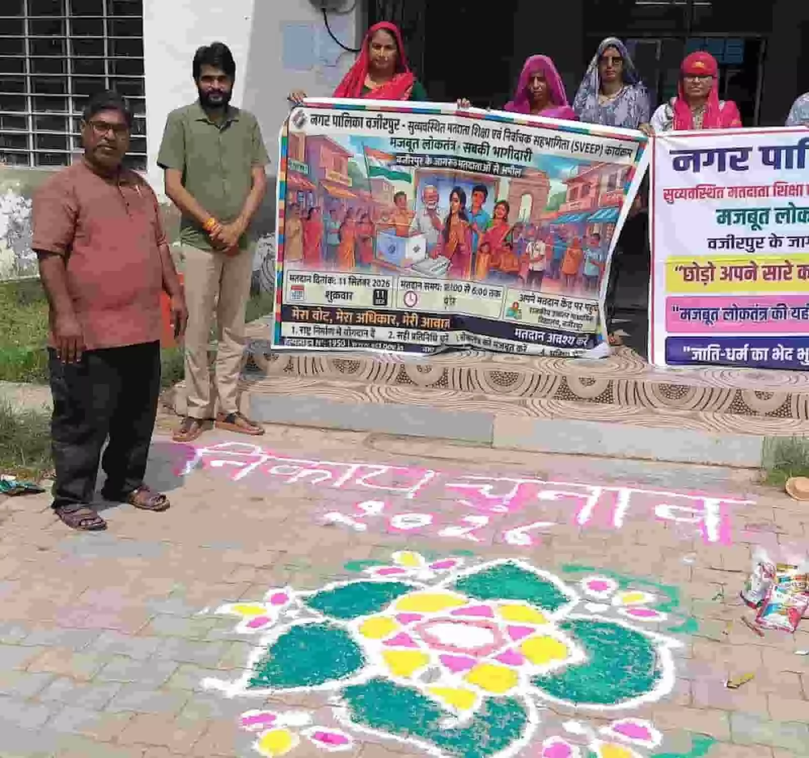 तस्वीर में बाईं ओर दो पुरुष और पीछे आंगनबाड़ी कार्यकर्ता स्वीप बैनर थामे जमीन पर बनी रंगोली के पास खड़े हैं। तस्वीर में बाईं ओर दो पुरुष और पीछे आंगनबाड़ी कार्यकर्ता स्वीप बैनर थामे जमीन पर बनी रंगोली के पास खड़े हैं।