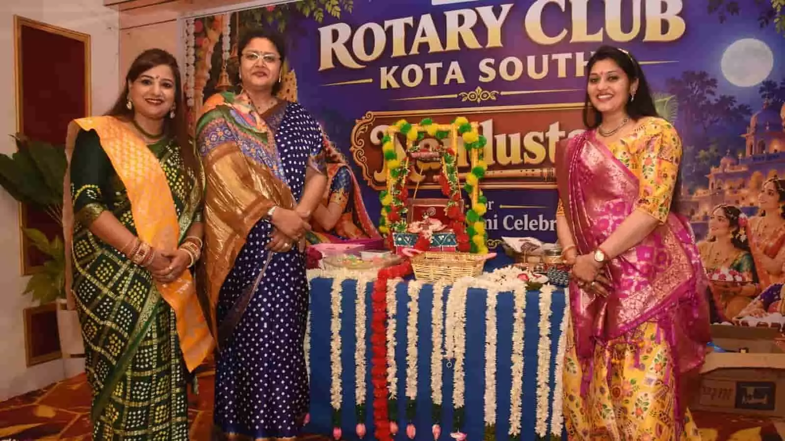 तस्वीर में पारंपरिक साड़ी पहने तीन महिलाएं सजे हुए मेज के पीछे खड़ी हैं। तस्वीर में पारंपरिक साड़ी पहने तीन महिलाएं सजे हुए मेज के पीछे खड़ी हैं।