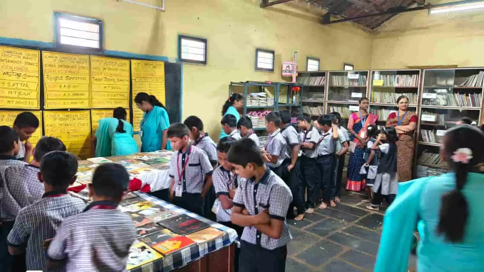विद्यालय के पुस्तकालय कक्ष में मेज पर सजी पुस्तकें देखते छात्र और पीछे खड़ी शिक्षिकाएं। विद्यालय के पुस्तकालय कक्ष में मेज पर सजी पुस्तकें देखते छात्र और पीछे खड़ी शिक्षिकाएं।
