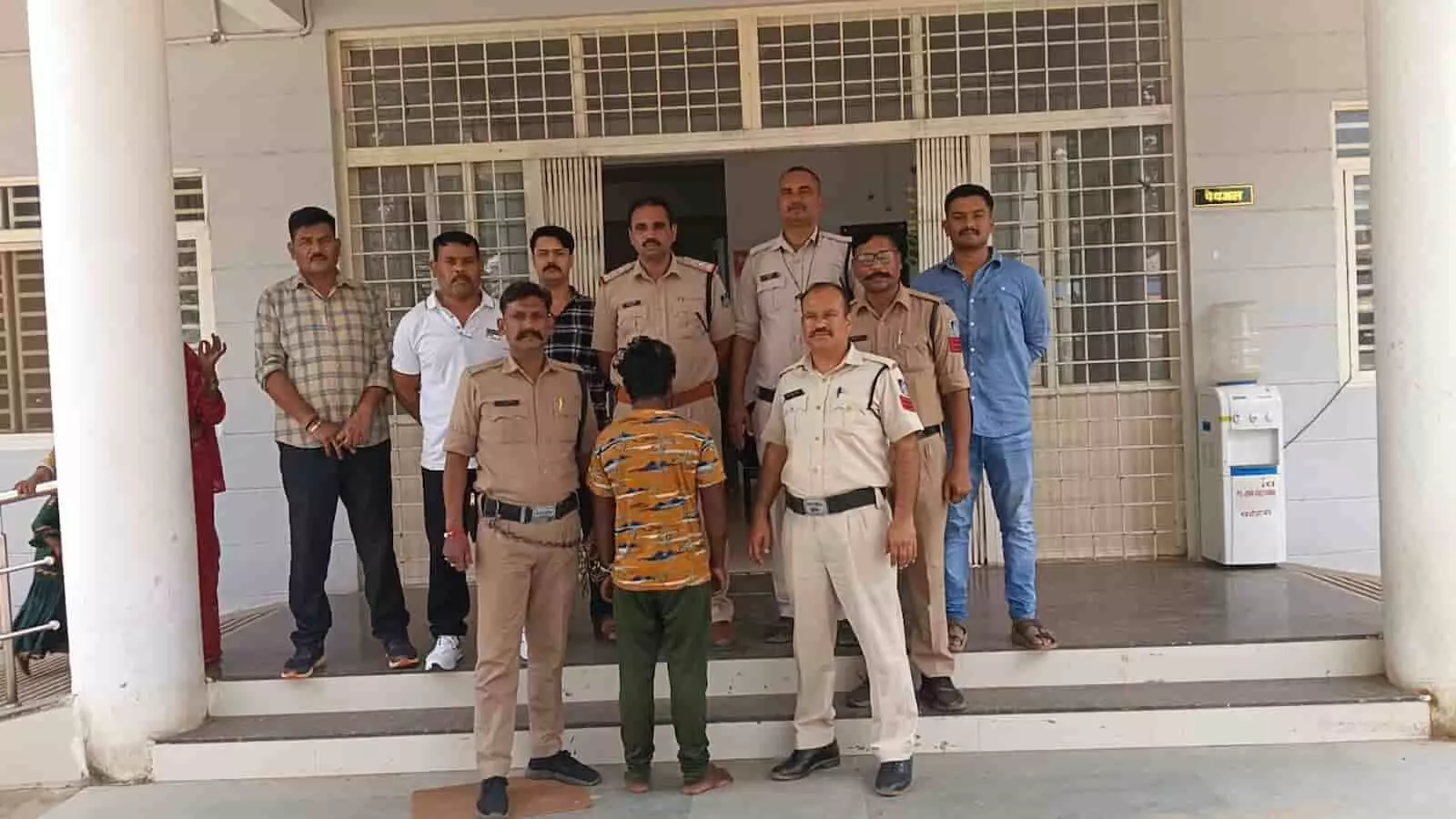 तस्वीर में दिख रही पुलिस टीम आरोपी को हिरासत में लेकर इमारत के बाहर खड़ी है। तस्वीर में दिख रही पुलिस टीम आरोपी को हिरासत में लेकर इमारत के बाहर खड़ी है।