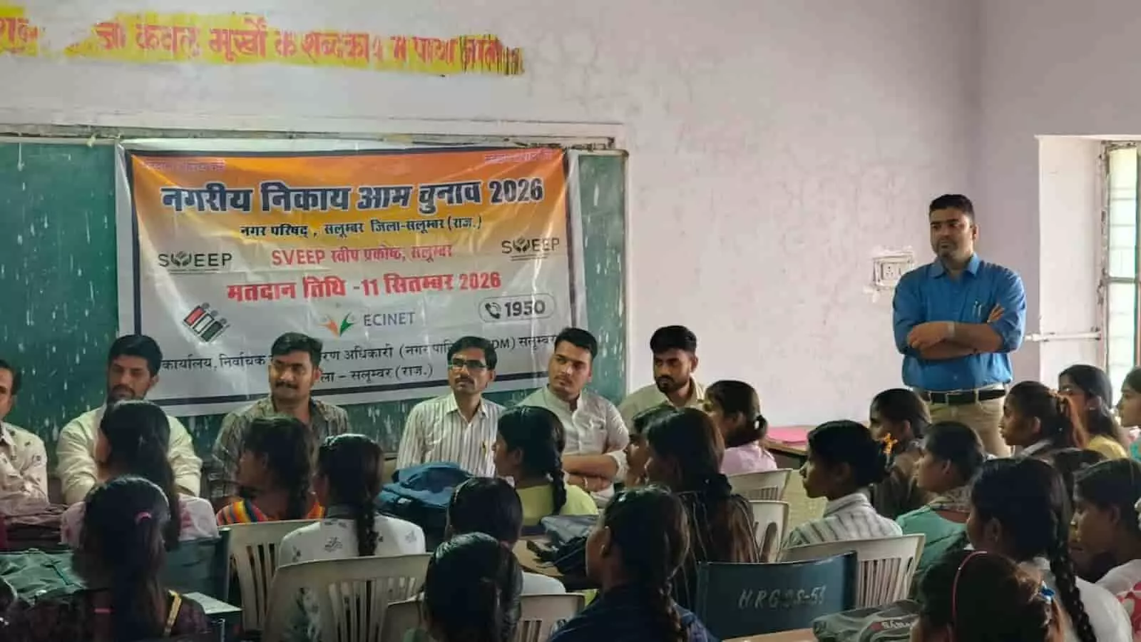 तस्वीर में दिख रही एक कमरे में चुनाव अधिकारी बैनर के पास लोगों को संबोधित कर रहे हैं। तस्वीर में दिख रही एक कमरे में चुनाव अधिकारी बैनर के पास लोगों को संबोधित कर रहे हैं।