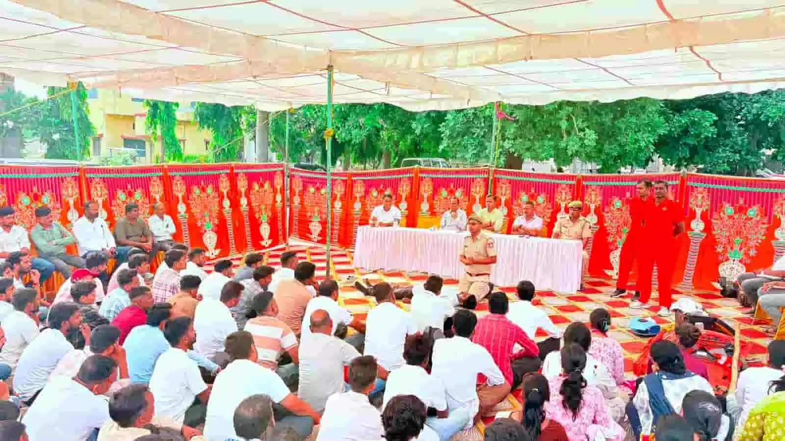 तस्वीर में दिख रही एक सभा में अधिकारी मंच पर बैठे हैं और सुरक्षाकर्मी स्वयंसेवकों को जानकारी दे रहे हैं। तस्वीर में दिख रही एक सभा में अधिकारी मंच पर बैठे हैं और सुरक्षाकर्मी स्वयंसेवकों को जानकारी दे रहे हैं।