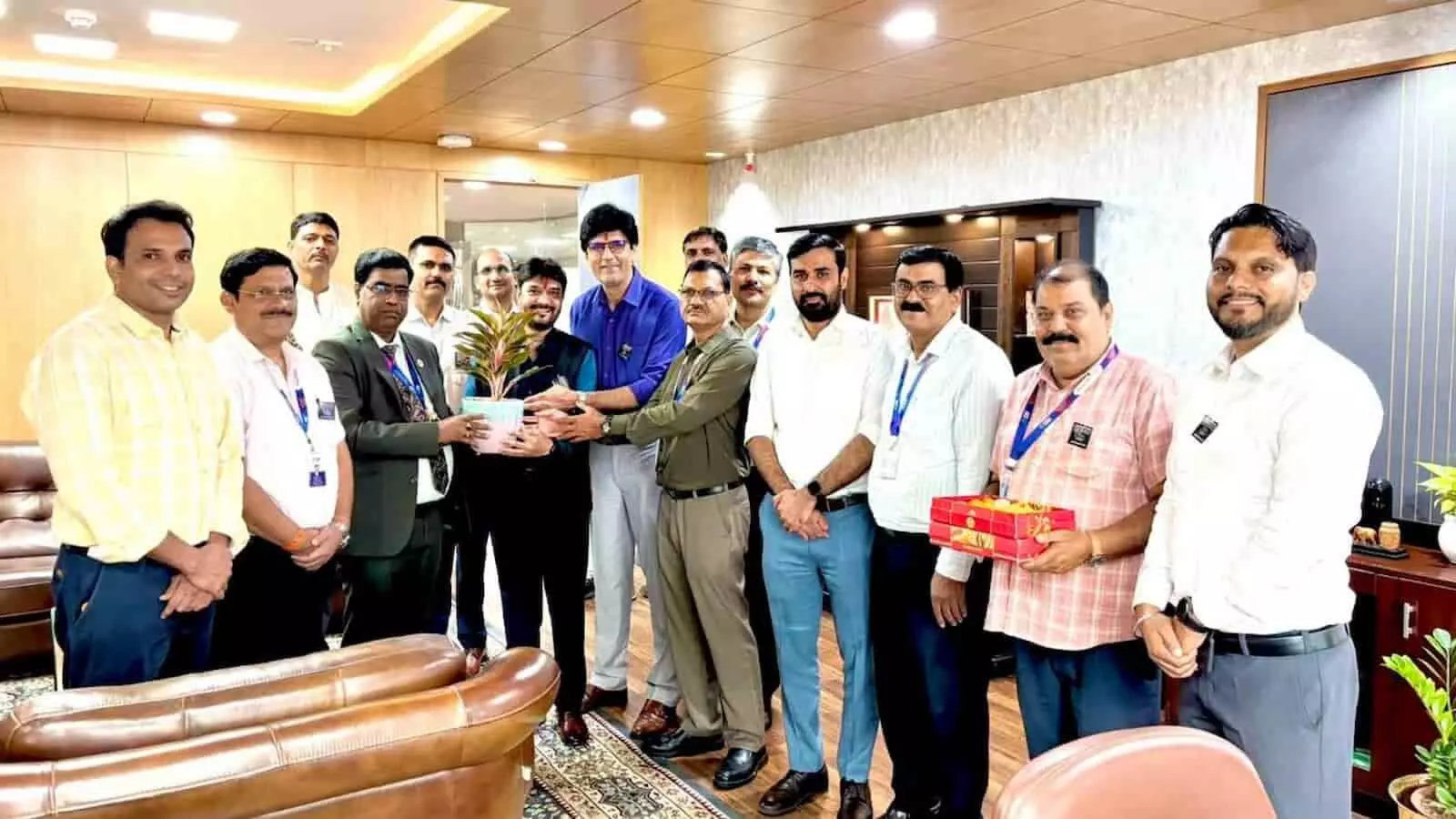 तस्वीर में बैंक के कार्यालय के भीतर खड़े कई अधिकारी, एक पौधे का गमला और मिठाई का डिब्बा थामे हुए दिखाई दे रहे हैं। तस्वीर में बैंक के कार्यालय के भीतर खड़े कई अधिकारी, एक पौधे का गमला और मिठाई का डिब्बा थामे हुए दिखाई दे रहे हैं।