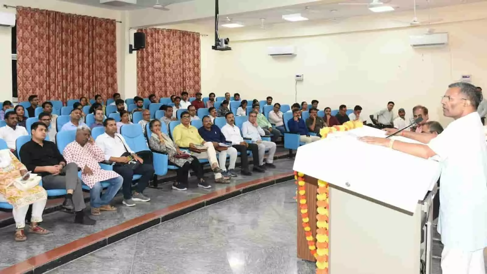 तस्वीर में एक व्यक्ति पोडियम के पास खड़े होकर भाषण दे रहा है और सामने सभागार में कई लोग बैठे हैं। तस्वीर में एक व्यक्ति पोडियम के पास खड़े होकर भाषण दे रहा है और सामने सभागार में कई लोग बैठे हैं।