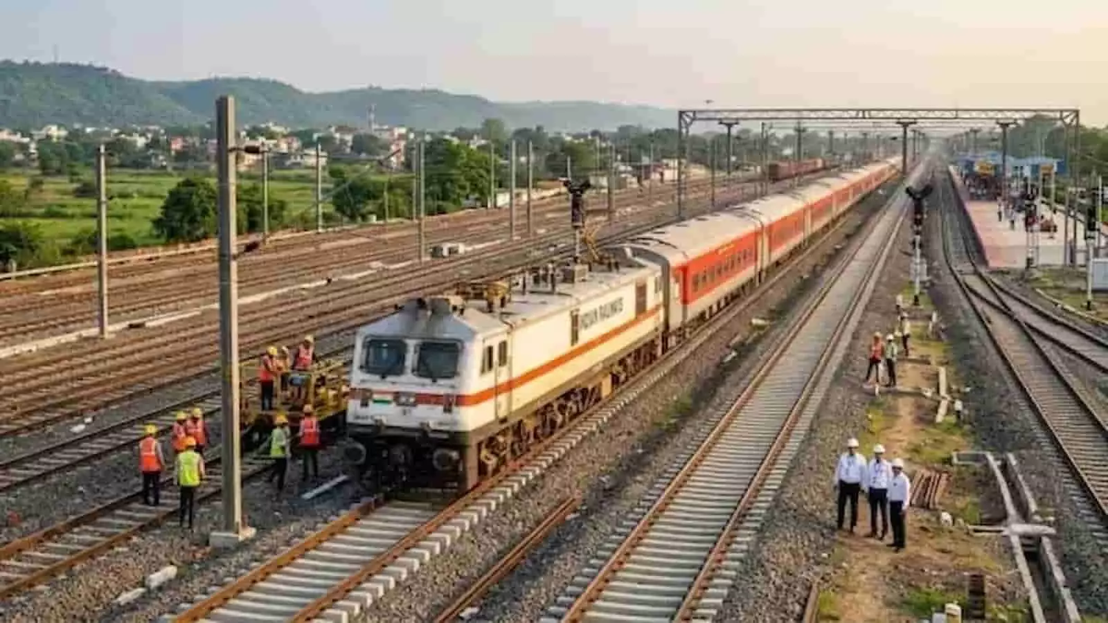 तस्वीर में दिख रही रेलवे पटरियों पर ट्रेन चलती हुई और पास में सुरक्षा जैकेट पहने रेल कर्मचारी काम करते हुए। तस्वीर में दिख रही रेलवे पटरियों पर ट्रेन चलती हुई और पास में सुरक्षा जैकेट पहने रेल कर्मचारी काम करते हुए।