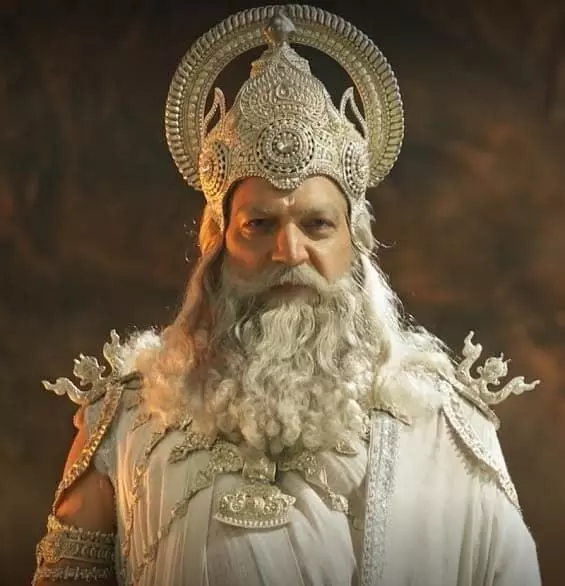 तस्वीर में सफेद लंबी दाढ़ी और भव्य मुकुट पहने भीष्म पितामह के रूप में अभिनेता का ऊपरी हिस्सा दिखाई दे रहा है। तस्वीर में सफेद लंबी दाढ़ी और भव्य मुकुट पहने भीष्म पितामह के रूप में अभिनेता का ऊपरी हिस्सा दिखाई दे रहा है।
