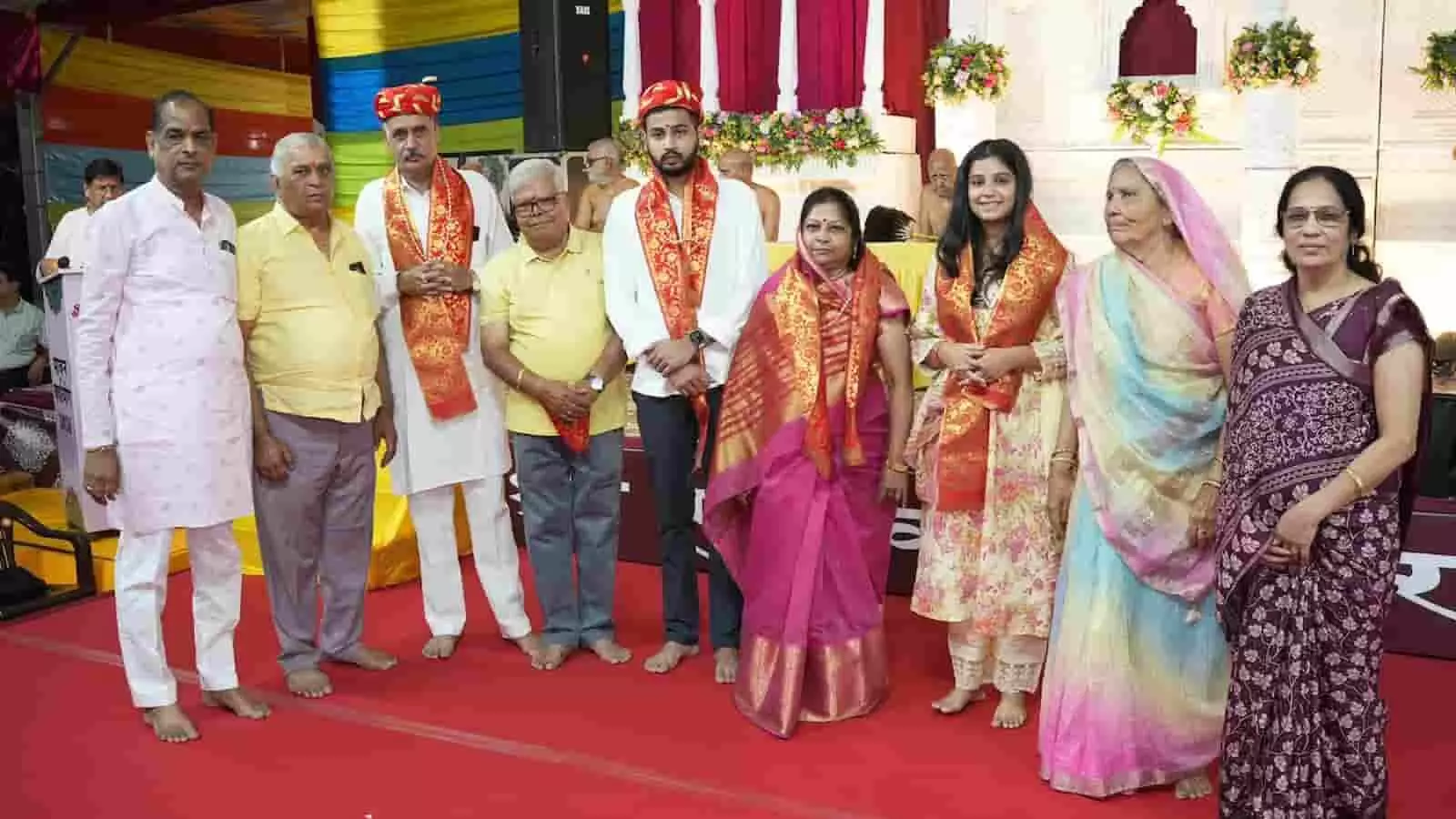 तस्वीर में बाएं से दाएं कई पुरुष और महिलाएं पारंपरिक वेशभूषा में मंच पर खड़े हैं, जिनमें कुछ लोगों ने गले में साफा पहना हुआ है। तस्वीर में बाएं से दाएं कई पुरुष और महिलाएं पारंपरिक वेशभूषा में मंच पर खड़े हैं, जिनमें कुछ लोगों ने गले में साफा पहना हुआ है।