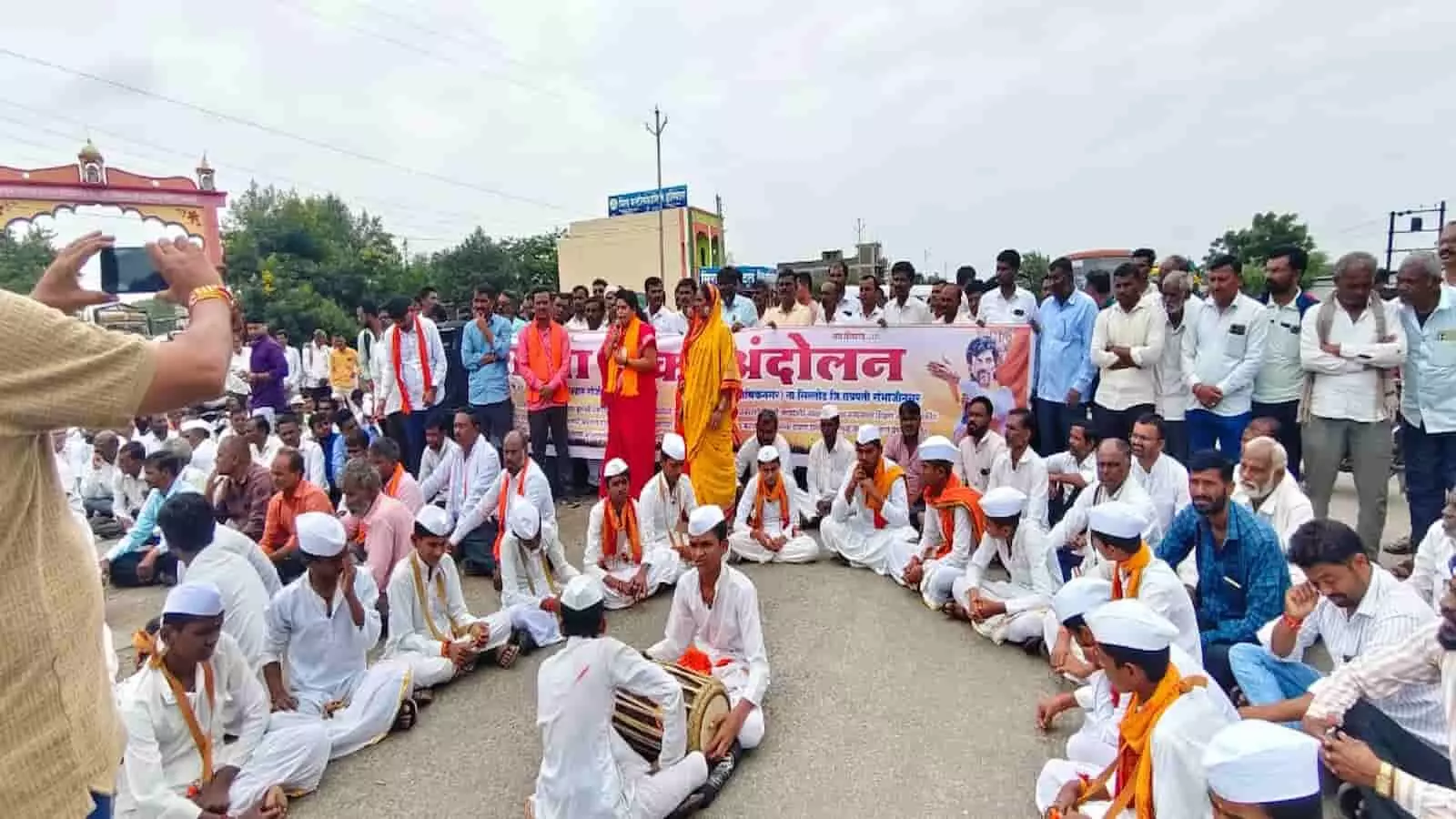 तस्वीर में बड़ी संख्या में लोग सड़क पर बैठे हुए हैं और पृष्ठभूमि में बैनर तथा एक भव्य प्रवेश द्वार दिखाई दे रहा है। तस्वीर में बड़ी संख्या में लोग सड़क पर बैठे हुए हैं और पृष्ठभूमि में बैनर तथा एक भव्य प्रवेश द्वार दिखाई दे रहा है।