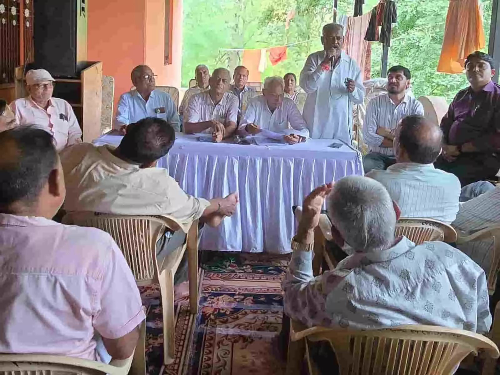 तस्वीर में मेज के पीछे खड़े बुजुर्ग व्यक्ति माइक पर बोलते हुए और बाकी लोग सुनते दिख रहे हैं। तस्वीर में मेज के पीछे खड़े बुजुर्ग व्यक्ति माइक पर बोलते हुए और बाकी लोग सुनते दिख रहे हैं।
