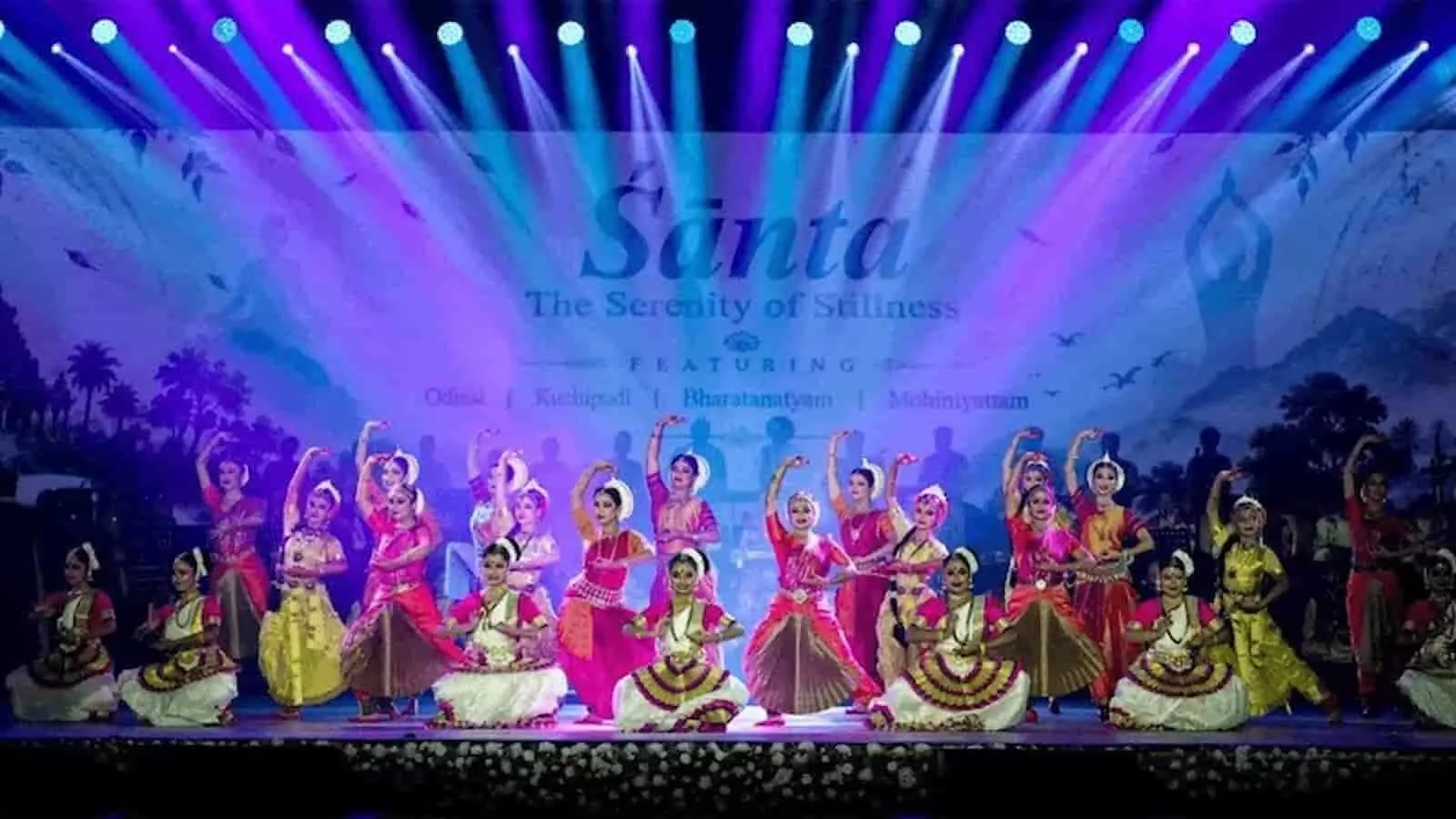 तस्वीर में रंग-बिरंगे प्रकाश के बीच मंच पर पारंपरिक नृत्य मुद्रा में कलाकारों का समूह दिख रहा है। तस्वीर में रंग-बिरंगे प्रकाश के बीच मंच पर पारंपरिक नृत्य मुद्रा में कलाकारों का समूह दिख रहा है।