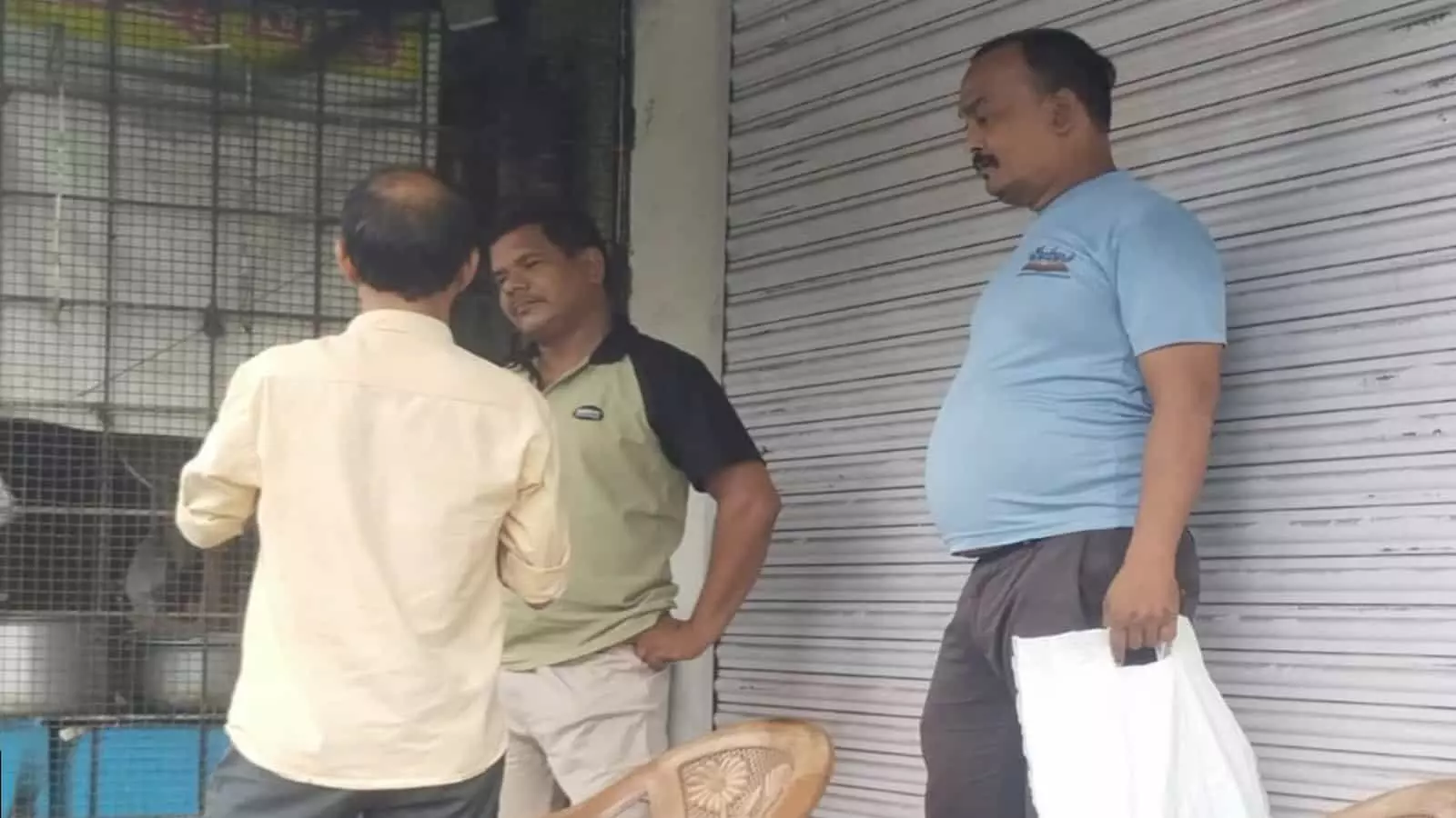 तस्वीर में एक बंद शटर वाली दुकान के बाहर प्लास्टिक की कुर्सियों के पास खड़े कुछ लोग दिखाई दे रहे हैं। तस्वीर में एक बंद शटर वाली दुकान के बाहर प्लास्टिक की कुर्सियों के पास खड़े कुछ लोग दिखाई दे रहे हैं।