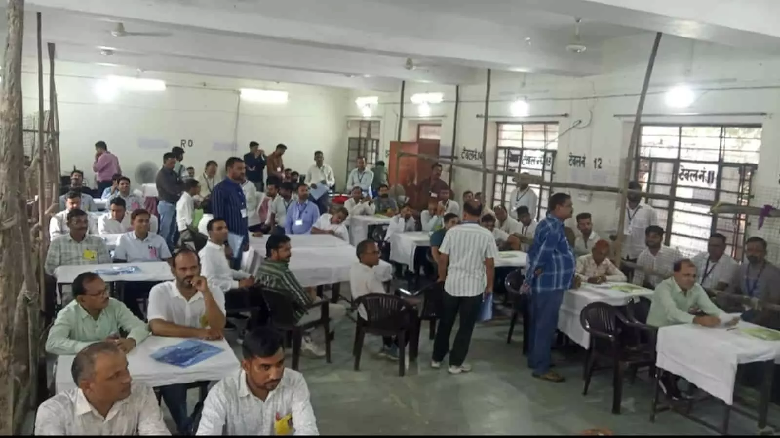 तस्वीर में बड़े हॉल के अंदर कई लोग टेबलों के पास खड़े और बैठे हुए दिखाई दे रहे हैं। तस्वीर में बड़े हॉल के अंदर कई लोग टेबलों के पास खड़े और बैठे हुए दिखाई दे रहे हैं।
