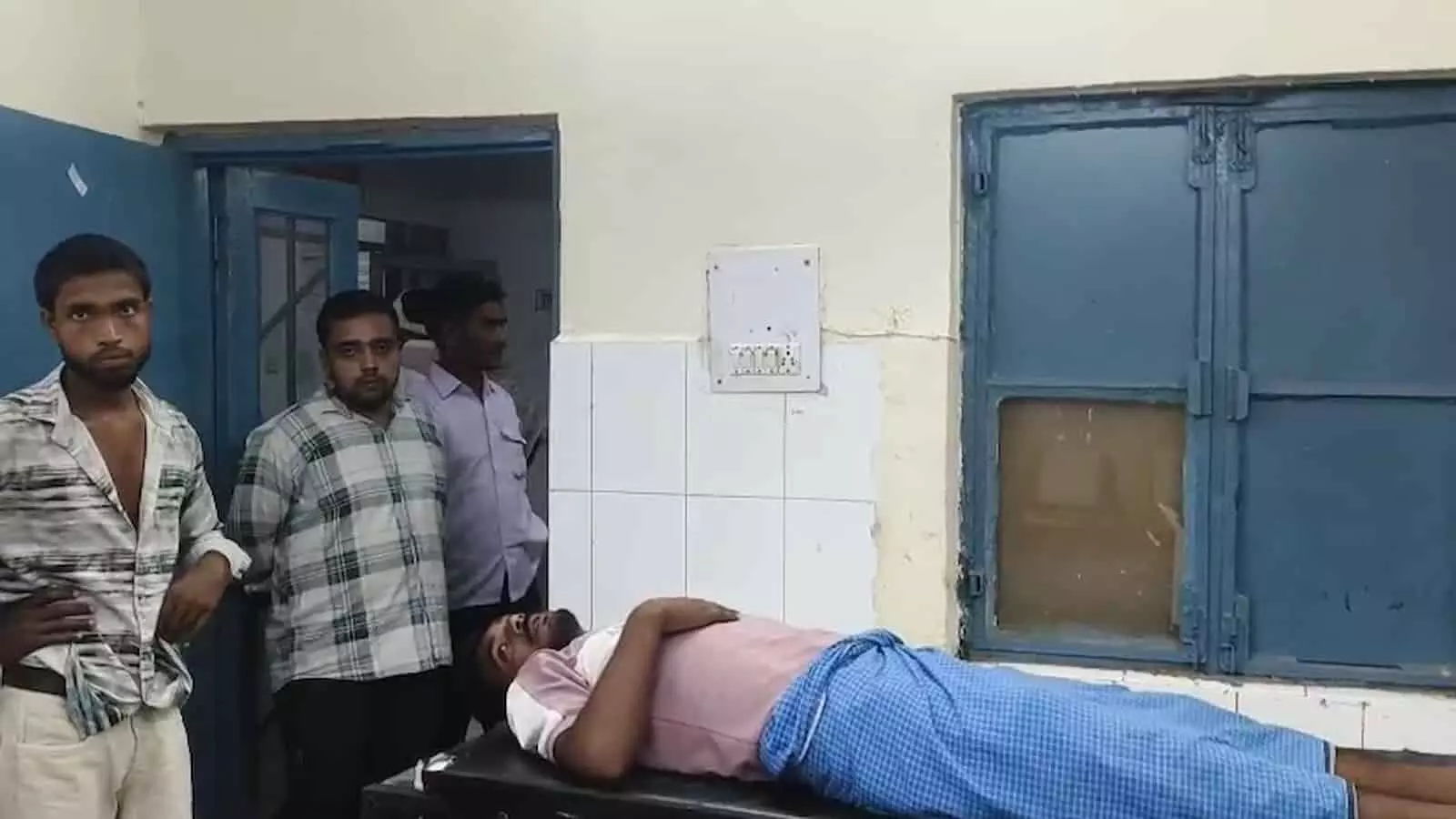 तस्वीर में अस्पताल के कमरे के अंदर एक घायल व्यक्ति लेटा है और तीन लोग खड़े हैं। तस्वीर में अस्पताल के कमरे के अंदर एक घायल व्यक्ति लेटा है और तीन लोग खड़े हैं।