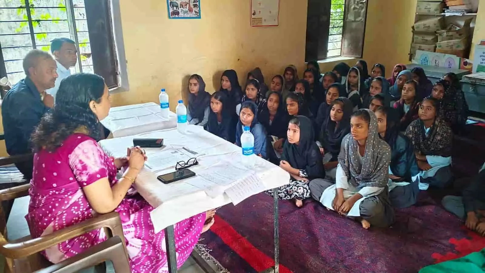 तस्वीर में एक कमरे के अंदर मेज के पास बैठी हुई महिला अधिकारी और सामने फर्श पर बैठी हुई कई स्कूली छात्राएं नजर आ रही हैं। तस्वीर में एक कमरे के अंदर मेज के पास बैठी हुई महिला अधिकारी और सामने फर्श पर बैठी हुई कई स्कूली छात्राएं नजर आ रही हैं।