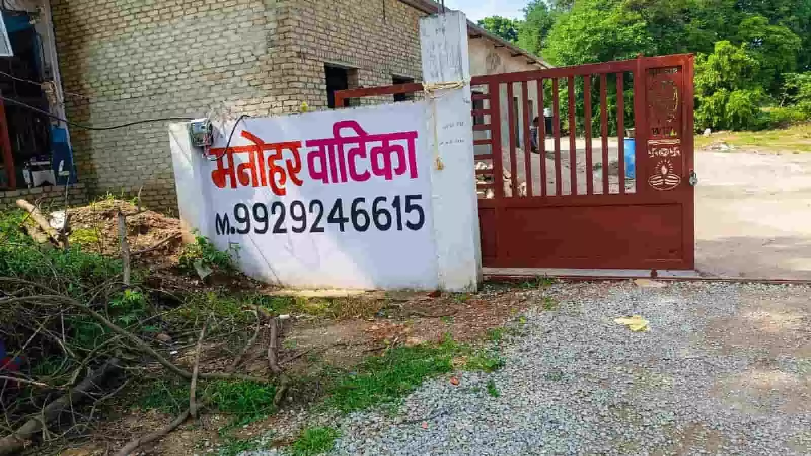 तस्वीर में दिख रही सफेद दीवार पर मनोहर वाटिका लिखा है और पास में लाल गेट है। तस्वीर में दिख रही सफेद दीवार पर मनोहर वाटिका लिखा है और पास में लाल गेट है।